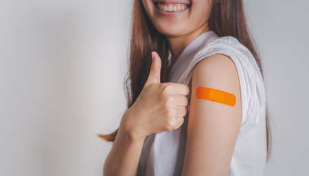 Asian woman showing thumb up gesture and shows off an orange bandage after receiving the vaccine.