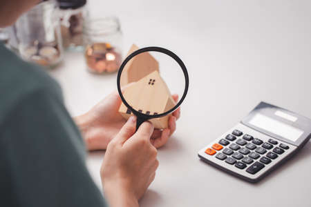 Woman's hand holding a magnifying glass looks at a model of a wooden house on a table with a...