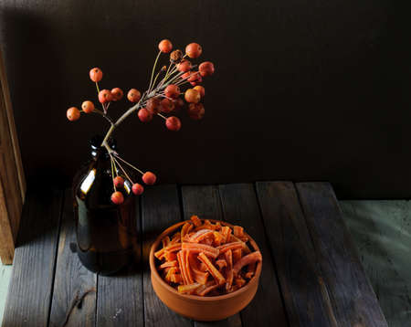 Carrot chips in ceramic bowl and dark glass bottle with apple tree branch with small fruits on...