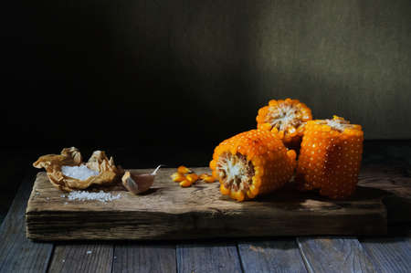 Baked corn ears with garlic and salt on narrow wooden board located on dark backgroundの写真素材