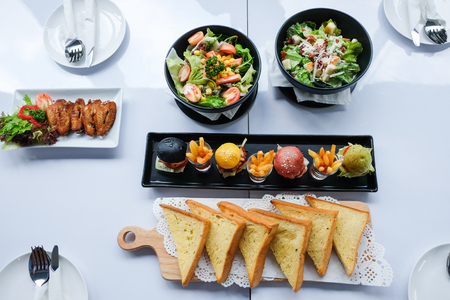 food course (burger, salads, garlic bread, chicken wings) on the white tableの写真素材