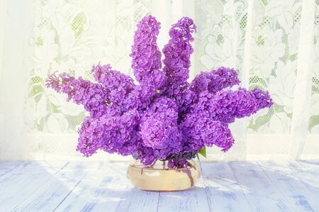 A bouquet of branches of lilac flowers purple, purple in a glass round vase on a wooden white table...
