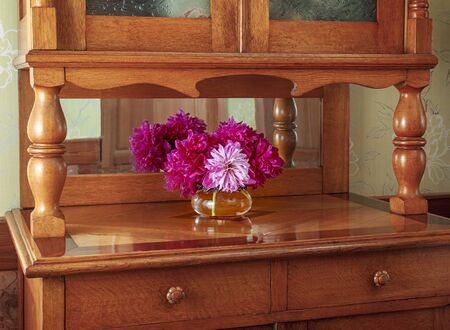 Vintage mahogany sideboard furniture with a vase with a bouquet of peony red and pink flowers that are reflected in the mirror.の写真素材