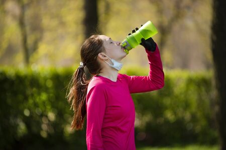 young girl drinks water after playing sports in the park. Girl in medical mask and gloves after quarantineの写真素材