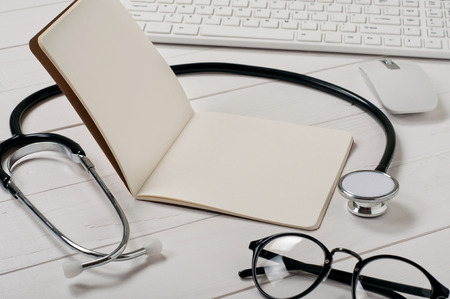 Open notebook with blank pages with stethoscope, keyboard, computer mouse and stylish sunglasses on a white desk close up.の写真素材