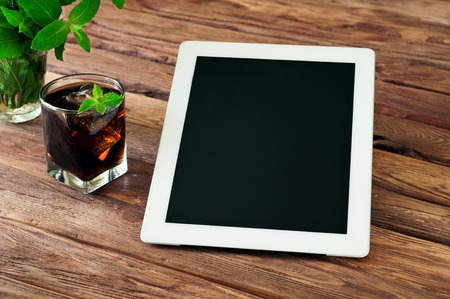 White tablet computer with a glass glass of cola with ice and mint on wooden background.