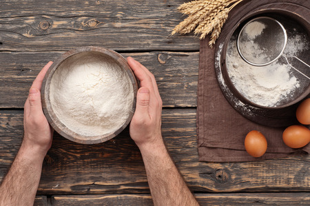 Baker holds a wooden bowl with flour powder on a dark wooden table in a bakery, top viewの写真素材