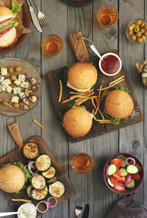 Different food on a wooden table, burgers, fries, grilled eggplant, salad, shiitake mushrooms, pickles, sauces, roquefort cheese, olives and light beer, top view.の写真素材