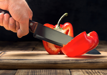 Man's hand with a kitchen knife cuts red pepper on a wooden cutting board on a black backgroundの写真素材
