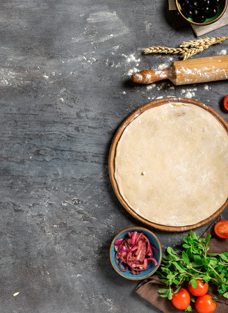 Raw pizza dough with ingredients for preparing vegetarian pizza on the dark stone surface with border, top viewの写真素材