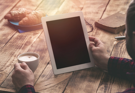 Close-up image of man holding digital tablet with copy space while sitting at the wooden table with...