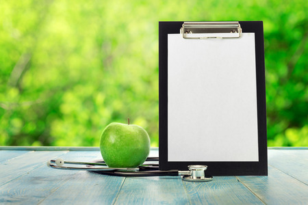 Clipboard with green apple and stethoscope on a blue wooden table against the background blur green leaves bokehの写真素材