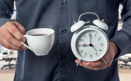 The office worker holds a cup of aromatic coffee and an alarm clock. Concept of morning coffee at workの写真素材