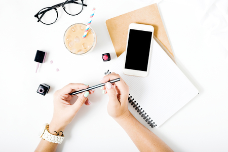 Home office workspace with woman hands, latte ice, notebook, spectacles, nail polish, lipstick and smartphone with blank screen on white background. Flat lay, top viewの写真素材