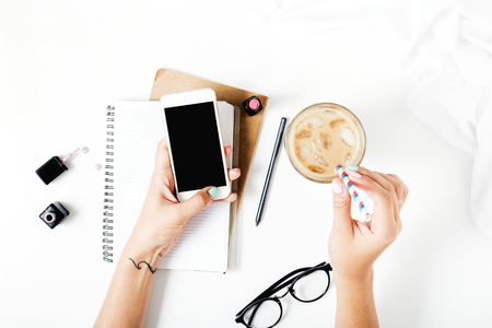 Home office workspace with smartphone in female hands, latte ice, notebook, spectacles, nail polish and lipstick. Flat lay, top viewの写真素材