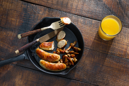 Top view fried chicken sausages with mushrooms in vintage frying pan on dark wooden table with glass of orange juiceの写真素材