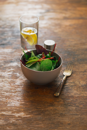 Vegetarian biodynamic food concept. Bowl of salad with spinach leaves and beet leaves on wooden table with water with lemonの写真素材