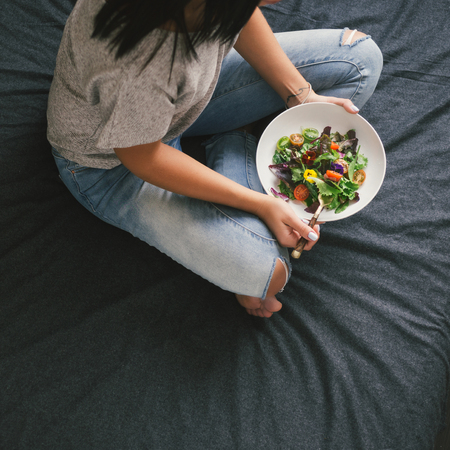Young woman eating salad on bed at homeの写真素材