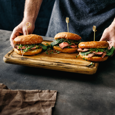 Male hands holding three burgers with grilled beef meat on wooden cutting board on dark backgroundの写真素材