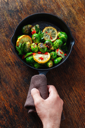 Male hand holding frying pan with fried Brussels sprouts on dark wooden background top viewの写真素材