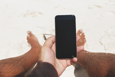 Male hands holding smartphone with blank screen sitting on the beach in summerの写真素材