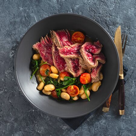 Dinner plate with beef steak with white beans, spinach and tomatoes on dark stone background top view with free coの写真素材