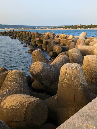 A series of large concrete tetrapods forming a coastal breakwater at a harbor, with a distant city skyline and calm sea under warm evening light.の写真素材