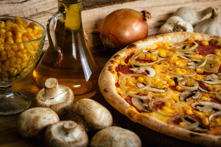 Whole pizza with sausages, corn and onion on wood table with ingredientsの写真素材