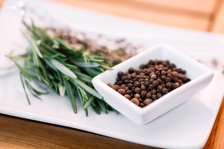 Rosemary with black pepper corns on a white plate on wood background