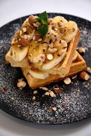 Sweet waffle with caramel and bananas sprinkled with hazelnuts and icing sugar on a white backgroundの写真素材