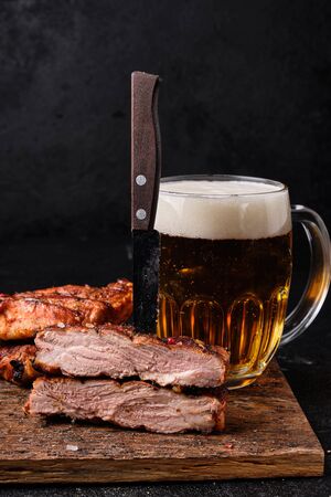 Cutted pork ribs and big glass of beer on cutting board on dark on concrete background. Knife stuck in meatの写真素材
