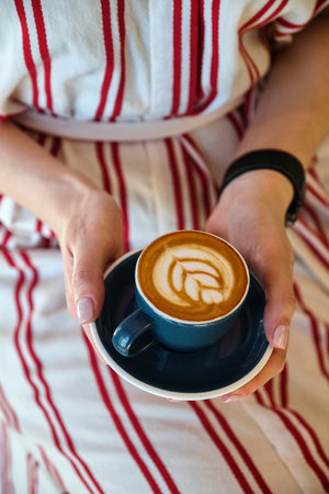 Girl in a light summer dress holds a cup of cappuccino with flower on top.