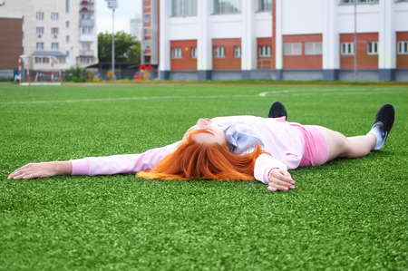 Beautiful sporty red-haired girl resting on grass after sport in the stadiumの写真素材