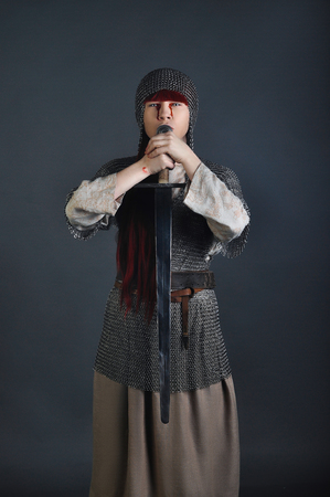 Medieval girl in chain mail with a sword with red hair stands on a gray background with blood on her hands. Studio shotの写真素材