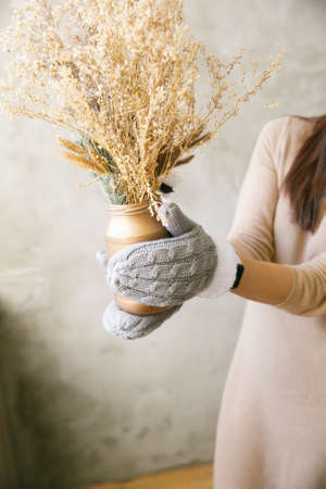 A girl in mittens holds a vase with dried flowers near the window in the morningの写真素材