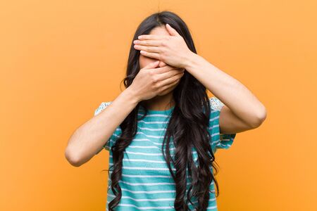young pretty hispanic woman covering face with both hands saying no to the camera! refusing pictures or forbidding photos against brown wallの写真素材