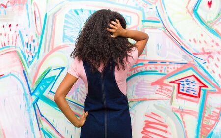 young pretty african american woman thinking or doubting, scratching head, feeling puzzled and confused, back or rear view against graffiti wallの写真素材