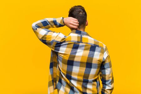 young hispanic man thinking or doubting, scratching head, feeling puzzled and confused, back or rear view against orange wallの写真素材