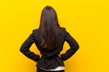 young  pretty woman feeling confused or full or doubts and questions, wondering, with hands on hips, rear view against orange wallの写真素材