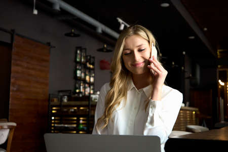 Cheerful blonde freelancer woman in white t-shirt talks on mobile phone with a clients while working...