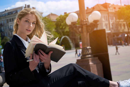 Attractive young blond woman reading a book on the go against the background of urban streetの写真素材