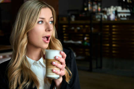 Young blonde woman drinking a cup of coffee scared and amazed with open mouth for surprise,...
