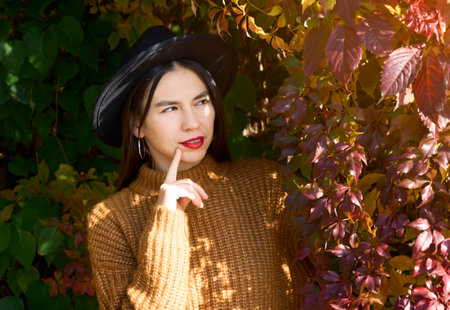 Selective focus of sweet dreamy young woman dressed brown outfit and hat. Finger on chin and thinking about autumn while standing in red and yellow leafs tree branches.の写真素材