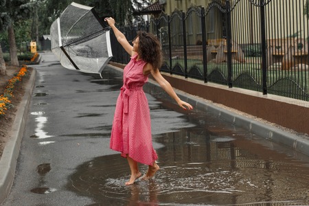 Young girl in a red dress with a transparent umbrella dancing in the rain standing in a puddle.