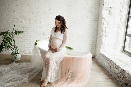 Portrait of beautiful pregnant girl sits near a bathtub with milk and flowers.