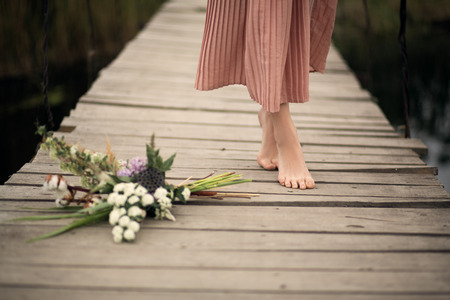 Close-up legs of beautiful tender girl in a peach-colored dress walking on a rural wooden bridge and bouquet of flowers on the floor.の写真素材