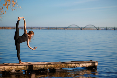 Young woman practicing yoga exercise at quiet wooden pier with city background. Sport and recreation in city rush.の写真素材
