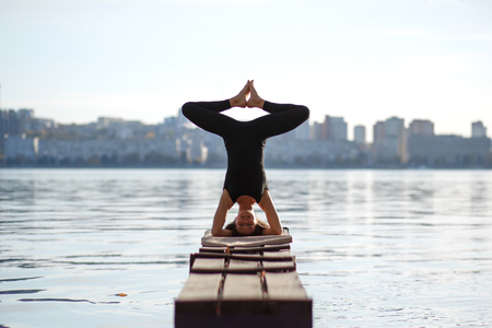 Young woman practicing yoga exercise at quiet wooden pier with city background. Sport and recreation in city rush.の写真素材
