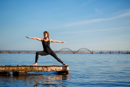 Young woman practicing yoga exercise at quiet wooden pier with city background. Sport and recreation in city rush.の写真素材