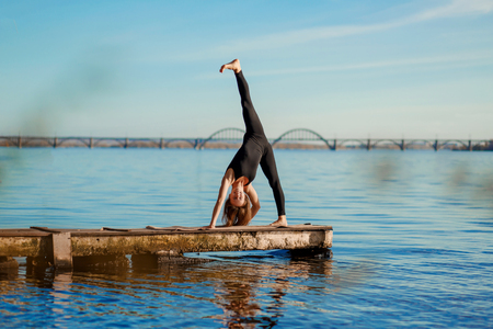 Young woman practicing yoga exercise at quiet wooden pier with city background. Sport and recreation in city rush.の写真素材